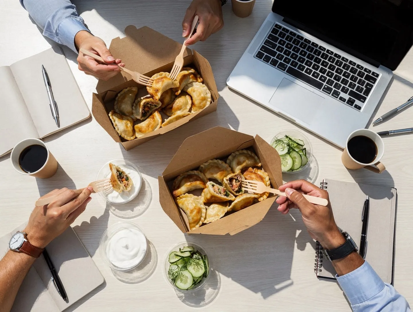 Offene Pappboxen mit Pierogi, Gurkensalat und Schmand auf einem Besprechungstisch zwischen Laptop und Notizbüchern