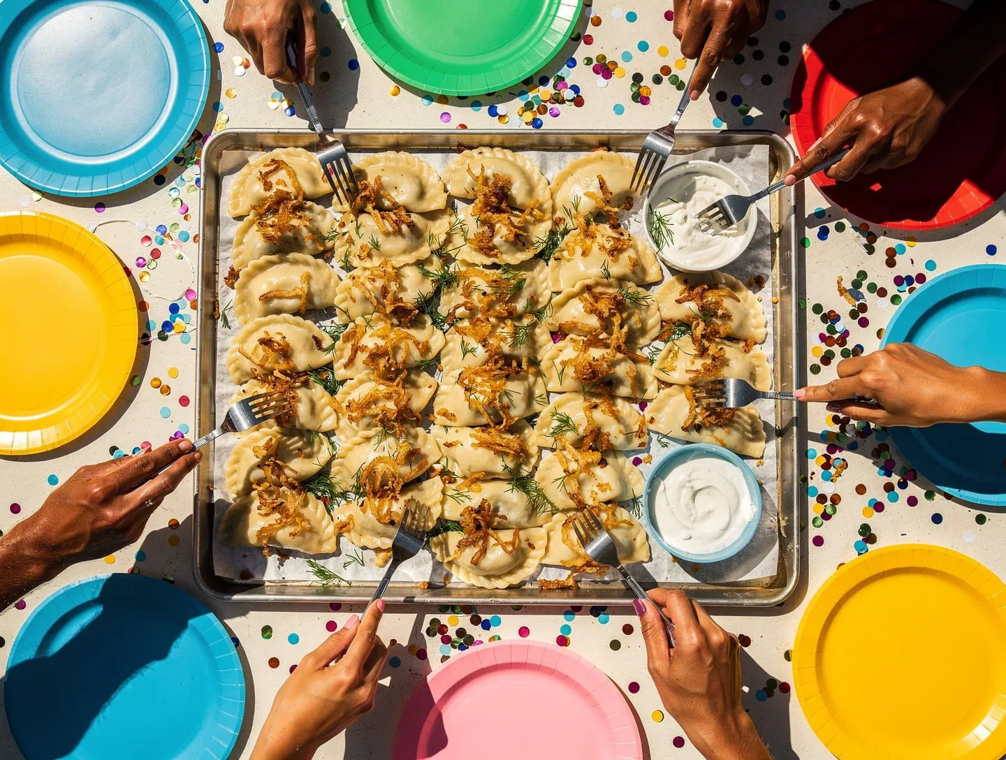Blech mit Reihen goldener und heller Pierogi auf einem bunten Geburtstagstisch, von allen Seiten greifen Hände zu