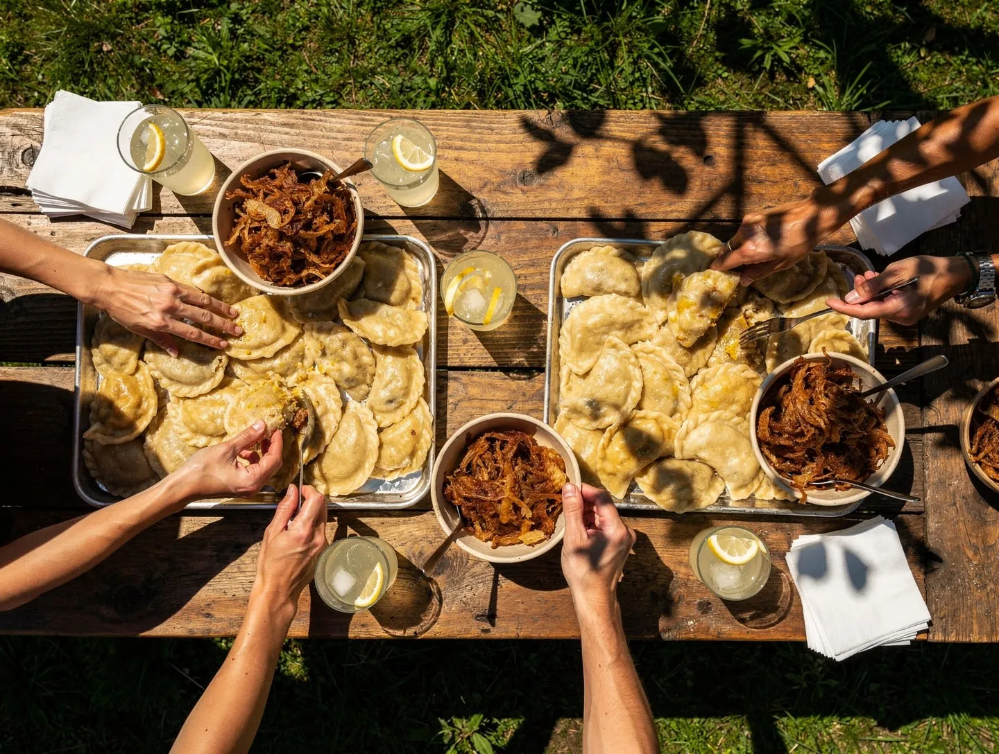 Langer Holztisch auf einer Wiese mit Blechen voller Pierogi, Schalen mit Zwiebeln und Gläsern Limonade